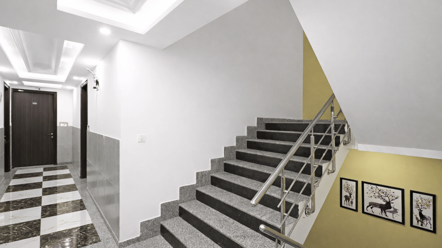 A view of the interior at Hotel Apple Park Delhi showing a stone staircase with a silver handrail, a yellow feature wall with framed artwork, and a chequered floor.
