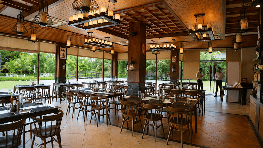 A restaurant seating area at Hotel Sonar Bangla, Sundarban featuring wooden dining tables, chairs, and light fixtures. 