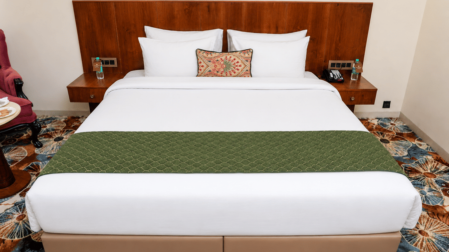 King bed with white bedding, decorative pillows, and a floral patterned carpet at Kadamb Kuteer, Vrindavan.
