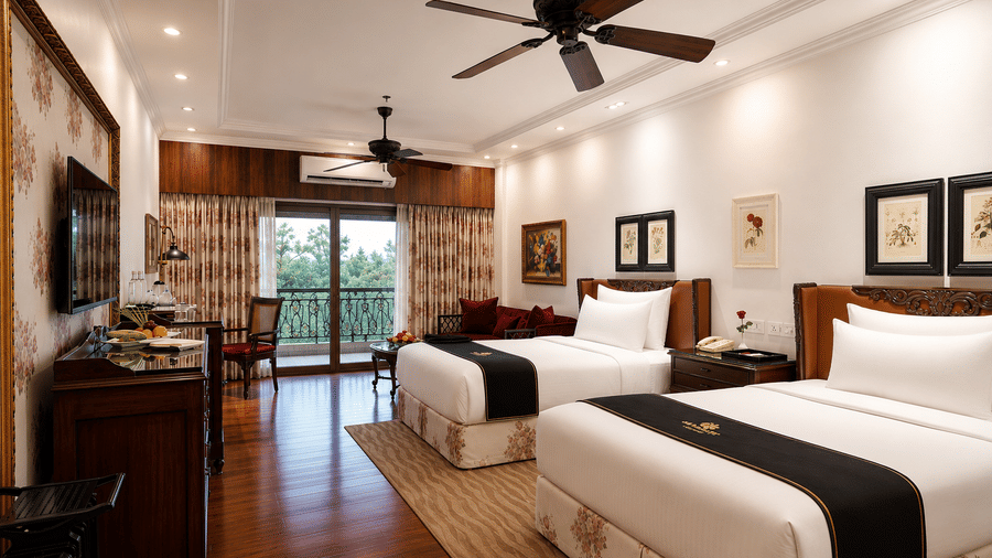 Deluxe Kanchenjunga room featuring twin beds inside a bedroom at MAYFAIR Himalayan Spa Resort Kalimpong. A ceiling fan is positioned above each bed. A television stands on a desk. A glass door opens onto a balcony with a view of foliage outside.