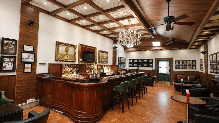 John's Tavern bar with a polished wooden counter, green leather stools, a crystal chandelier and framed vintage prints at MAYFAIR Himalayan Spa Resort, Kalimpong.