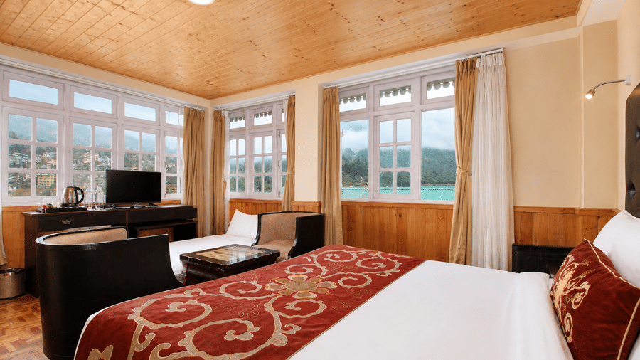 Deluxe room at Orsino Khangri Karpo Retreat & Spa, Lachung offers views through large windows. The space includes a wooden ceiling, a television, and a bed with patterned linens.