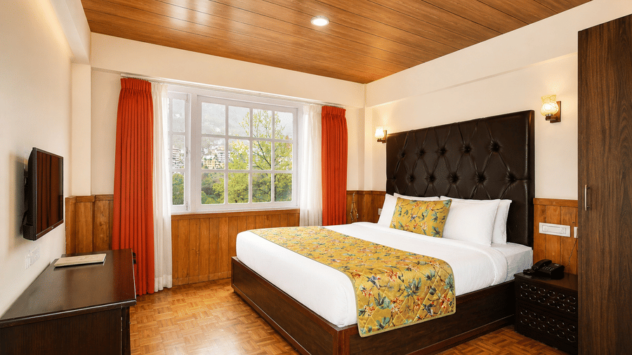 Deluxe Room at Orsino Khangri Karpo Retreat & Spa, Lachung, featuring a neatly-made bed, curtains framing a large window, wooden finishes and a television.