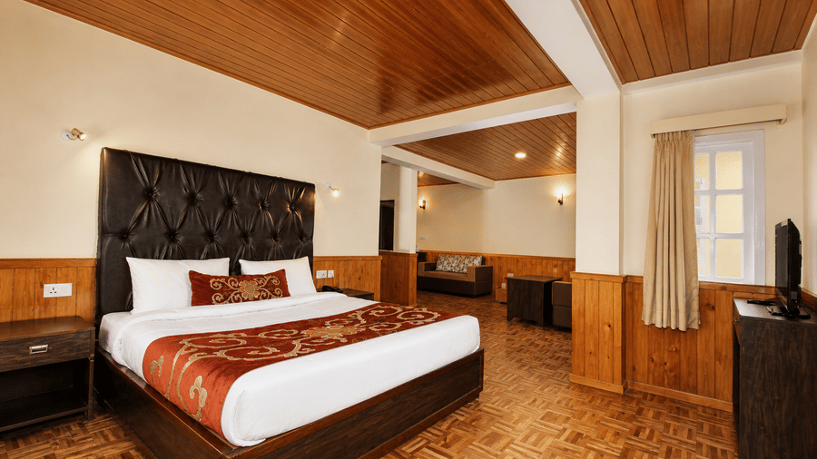 A bed sits in the centre of the Junior Suite at Orsino Khangri Karpo Retreat & Spa, Lachung. Wood panelling covers the walls and ceiling. A window and a television stand in the corner.