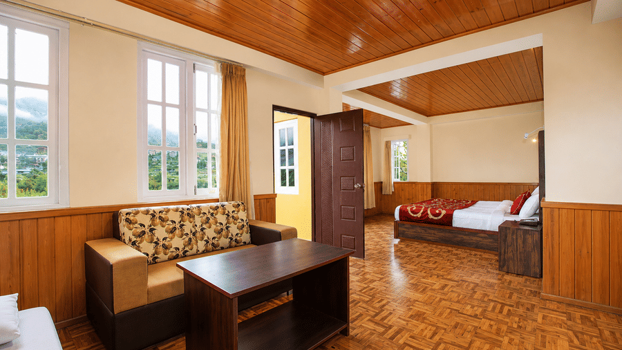 A sofa and a coffee table are placed in the Junior Suite at Orsino Khangri Karpo Retreat & Spa, Lachung. Windows look out from the wall. The bedroom area is visible through a doorway.