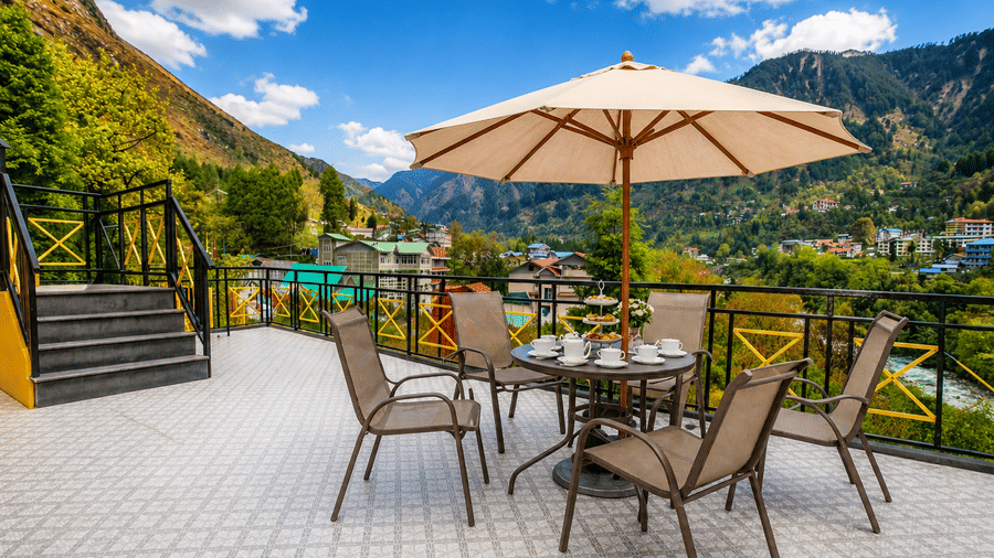 Opal bar at Orsino Khangri Karpo Retreat & Spa, Lachung featuring a central table with a sun umbrella and chairs sits on a paved area. Green mountains and a blue sky form the background.