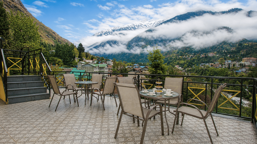 Opal bar at Orsino Khangri Karpo Retreat & Spa, Lachung featuring metal tables and chairs sit on a tiled floor. Mountains and clouds are visible beyond the black metal railing.