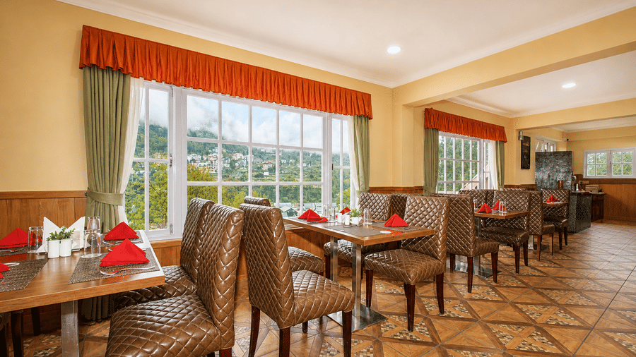 Orion restaurant at Orsino Khangri Karpo Retreat & Spa, Lachung featuring wooden tables and chairs with patterned upholstery stand on a tiled floor. Windows with curtains overlook the hills. Place settings include napkins and glassware.