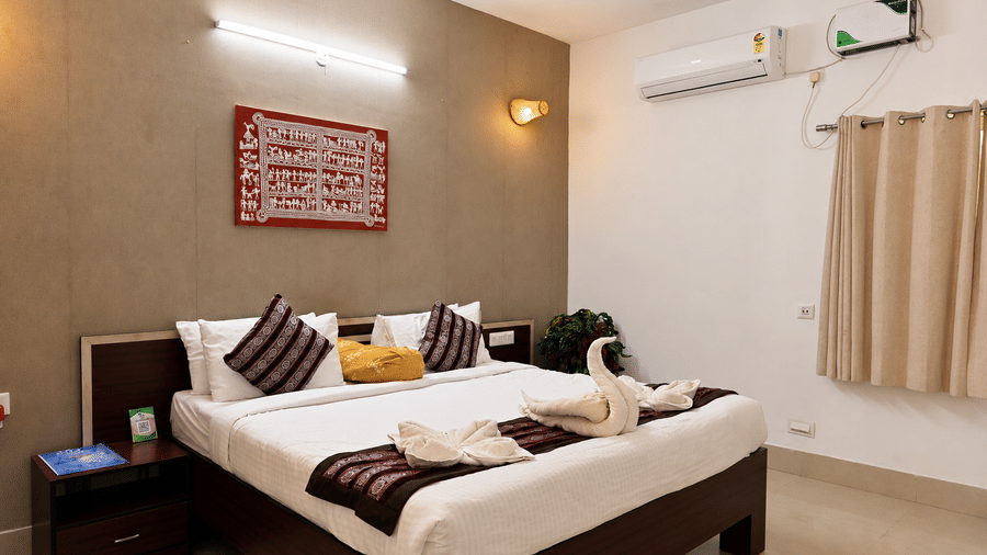 Hotel room with a bed dressed in white linen, swan-shaped towel art, patterned cushions, a wall-mounted artwork and a window with curtains.