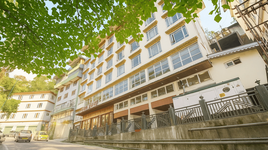 A multi-storey modern hotel building with large glass windows, built into a lush green hillside under a bright sky.