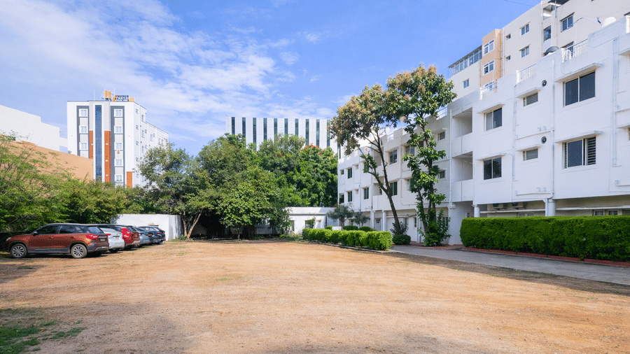 Exterior of The Pinnacle featuring a wide parking area bordered by multi-story buildings, a row of parked cars, and lush green trees under a bright sky.