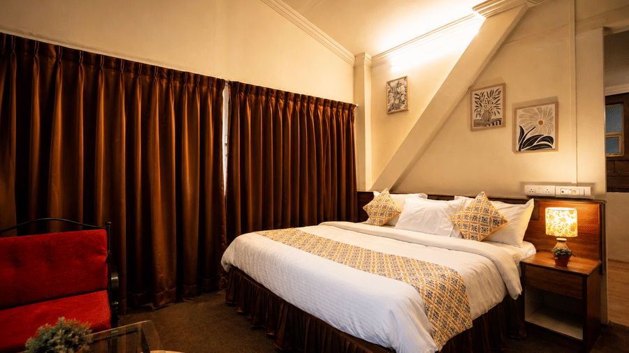 Warmly lit Wind Chalet bedroom with double bed, bedside lamp, framed art, red seat, and floor-to-ceiling brown drapes.