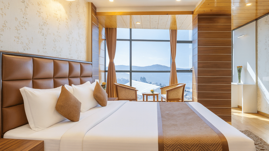 A spacious and well-lit bedroom with a large bed, seating area, and a window view at the Yashshree Jai Hotel & Banquet, Darjeeling.