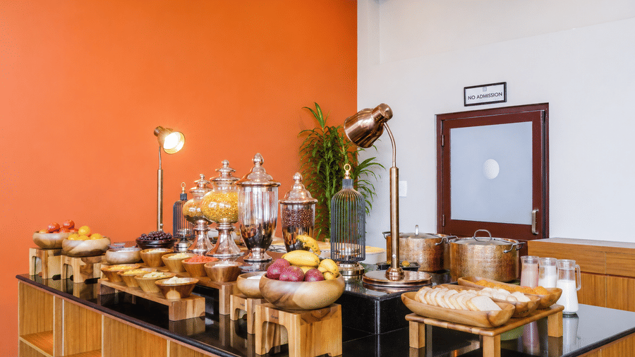 Breakfast buffet spread at Valmere Himalayan Hotel & Spa, Gangtok with fresh fruit bowls, cereal jars, bread, milk jugs, and a plant against an orange wall.