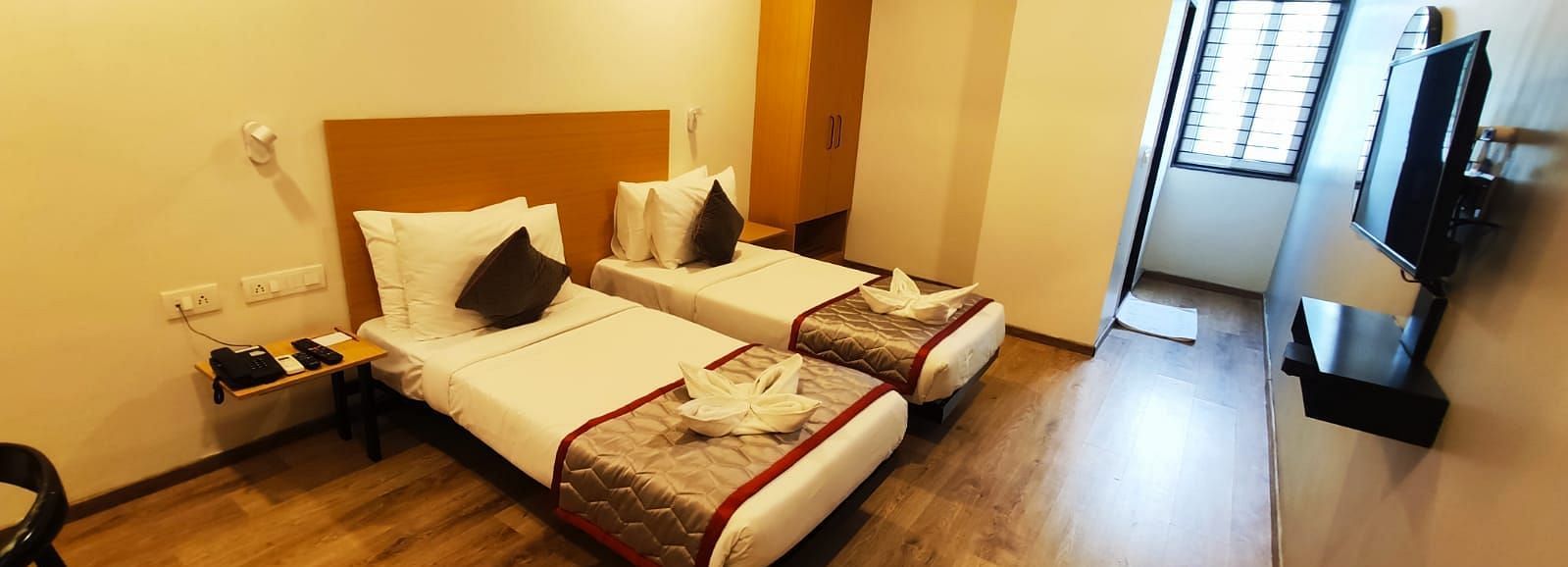Hotel bedroom with two single beds, wooden furniture, and a view through the window - Lords Eco Inn Belagavi
