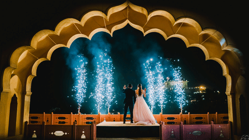 A couple stands on a raised platform under an arched structure with fireworks behind them at Tijara Fort-Palace - 19th Century, Alwar.