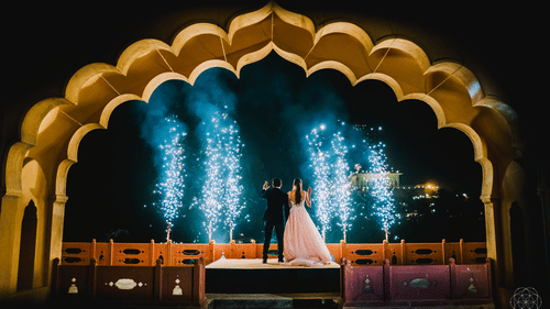 A couple stands on a raised platform under an arched structure with fireworks behind them at Tijara Fort-Palace - 19th Century, Alwar.