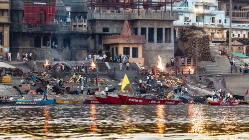 A river scene at Manikarnika Ghat in Varanasi, showing boats on the Ganges River and cremation ceremonies taking place on the shore.