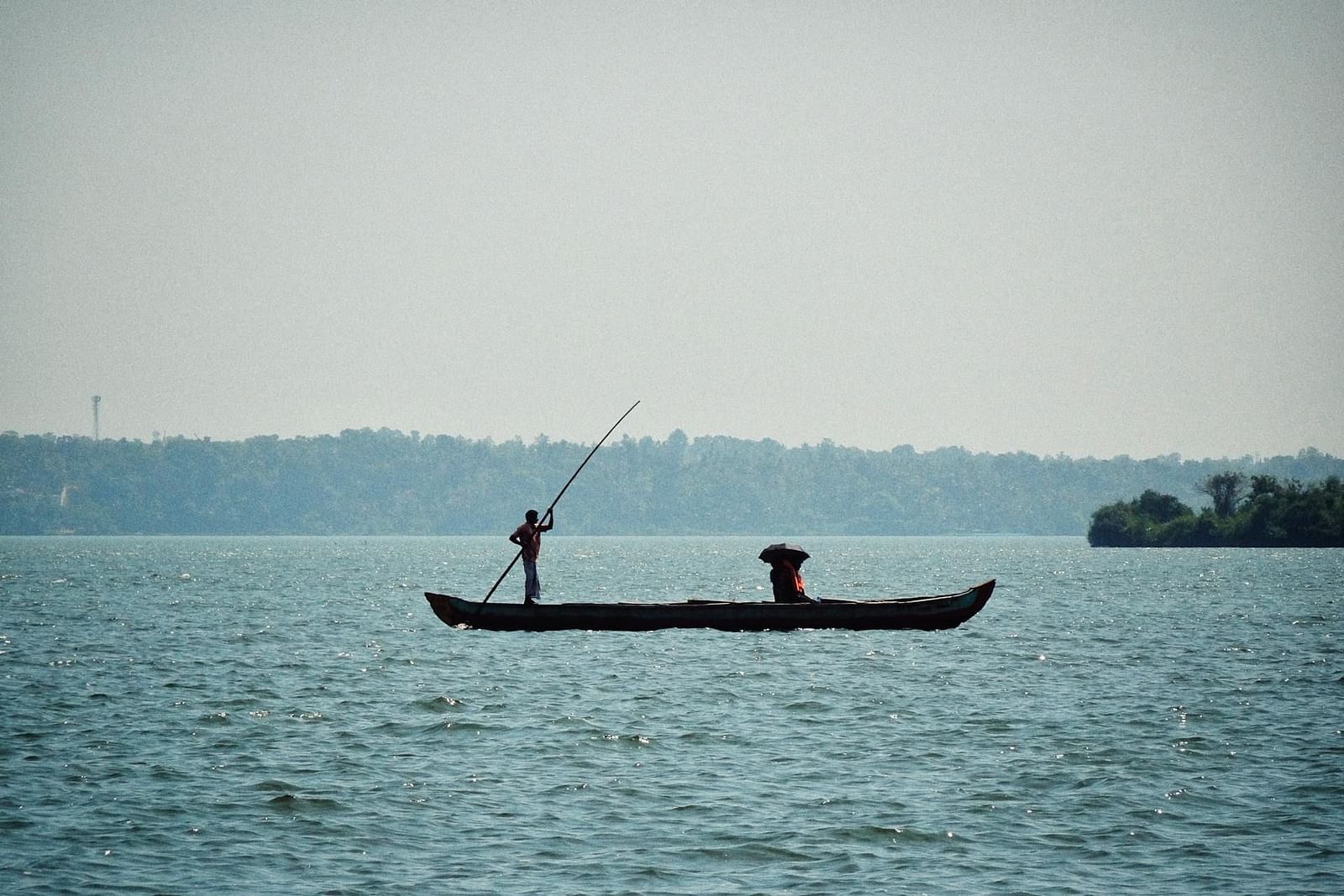 A person rows a small boat across a calm river under a clear sky.