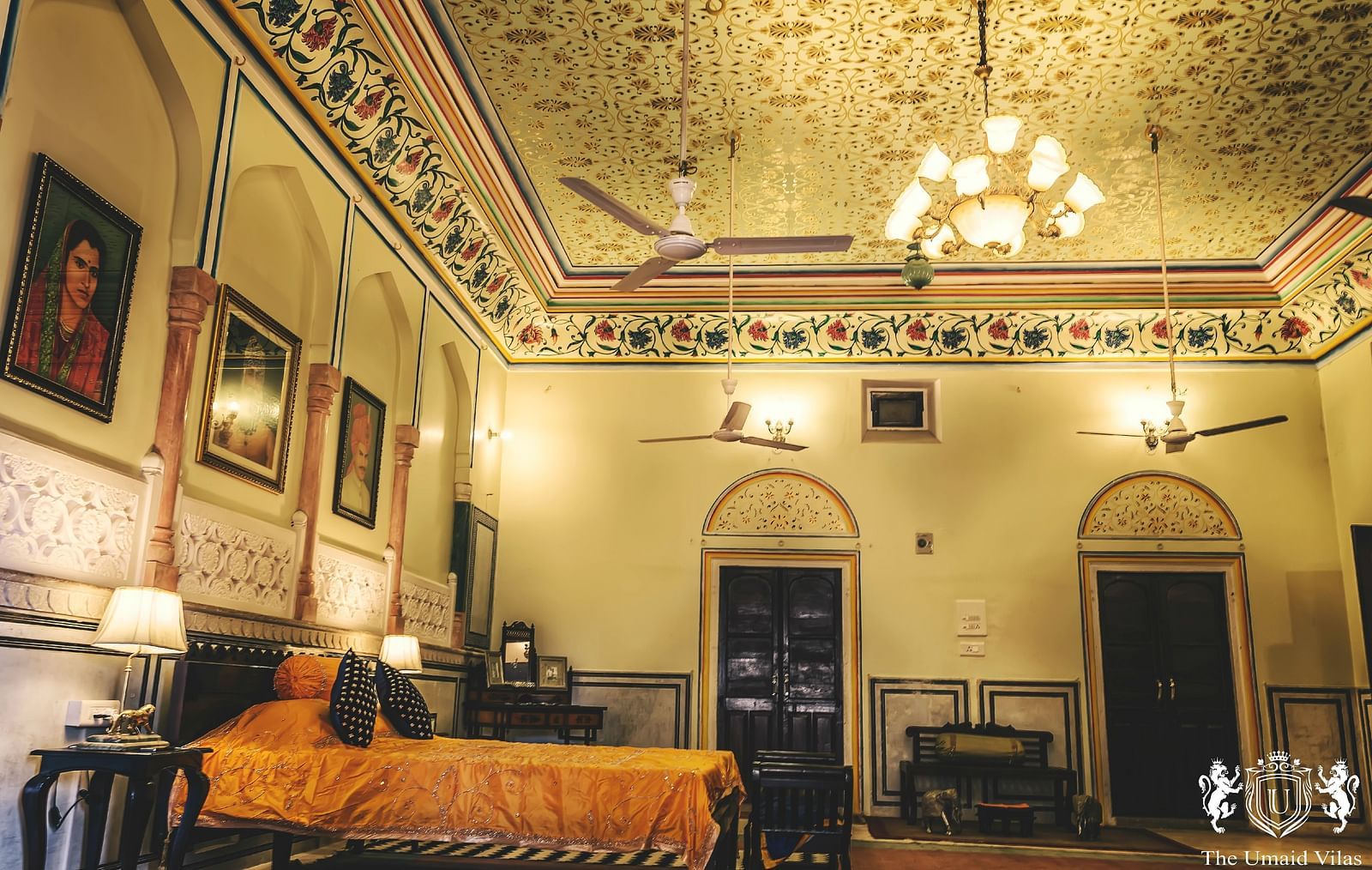 A spacious room at Umaid Vilas Jaipur with a patterned ceiling, multiple fans, and a bed with an orange throw.