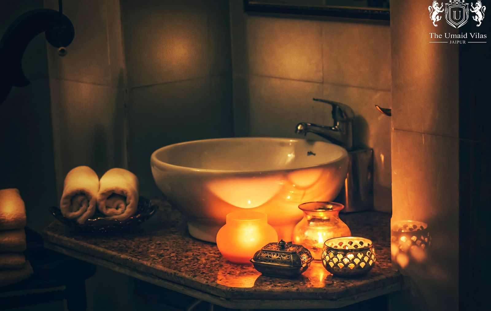 A dimly lit bathroom at Umaid Vilas Jaipur with a white sink, rolled towels, and several lit candles creating a warm, inviting atmosphere.