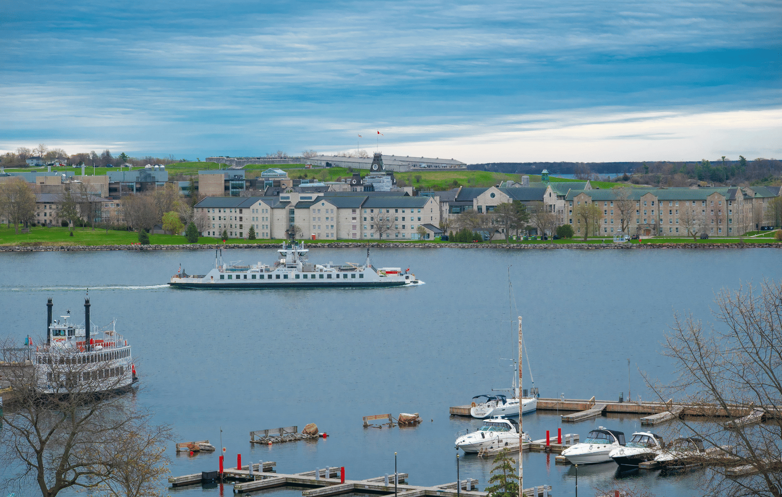 A scenic waterfront view featuring multiple small boats and vessels moored along a calm harbour, with a historic town skyline and architectural buildings stretching across the background shoreline.