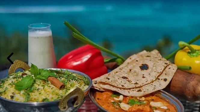 2 bowls of curry with flatbread (roti), vegetables, and a glass of buttermilk, blurred ocean in background | A Stone's Throw Away