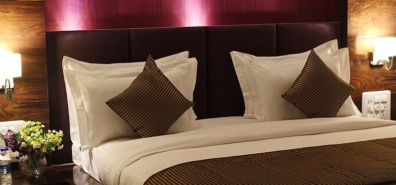 A warmly lit hotel room showcases a king size bed with a prominent dark headboard, crisp white linens, and decorative throw pillows.