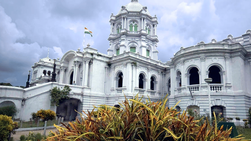Ujjayanta Palace heritage landmark in Agartala displaying white domed towers, arched balconies and Indian flag against a dramatic cloudy sky with garden foreground.