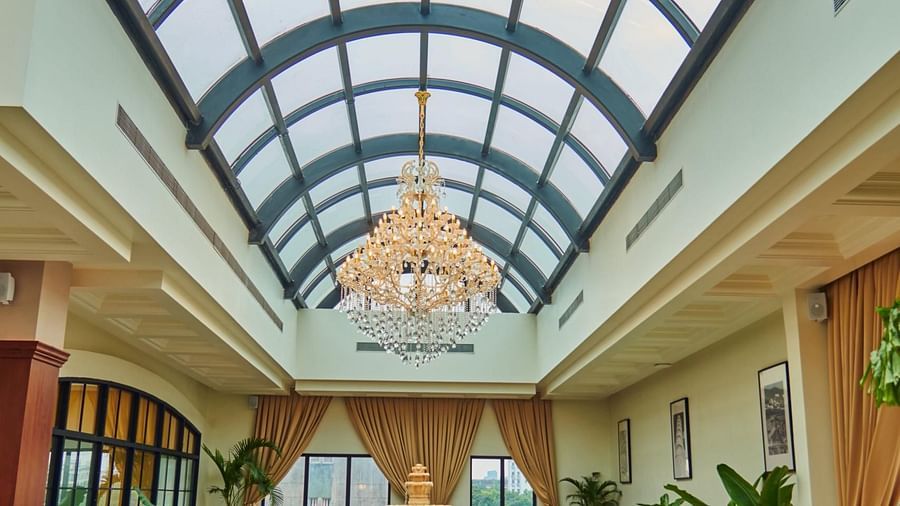 A spacious, elegant indoor Meli-Melo with a high arched glass ceiling, vibrant seating, a central fountain, chandeliers, large windows, and potted plants creating a bright, inviting atmosphere