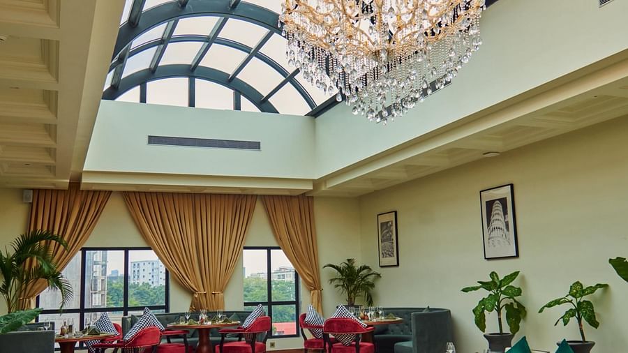 A spacious, elegant indoor Meli-Melo with a high arched glass ceiling, vibrant seating, a central fountain, chandeliers, large windows, and potted plants creating a bright, inviting atmosphere