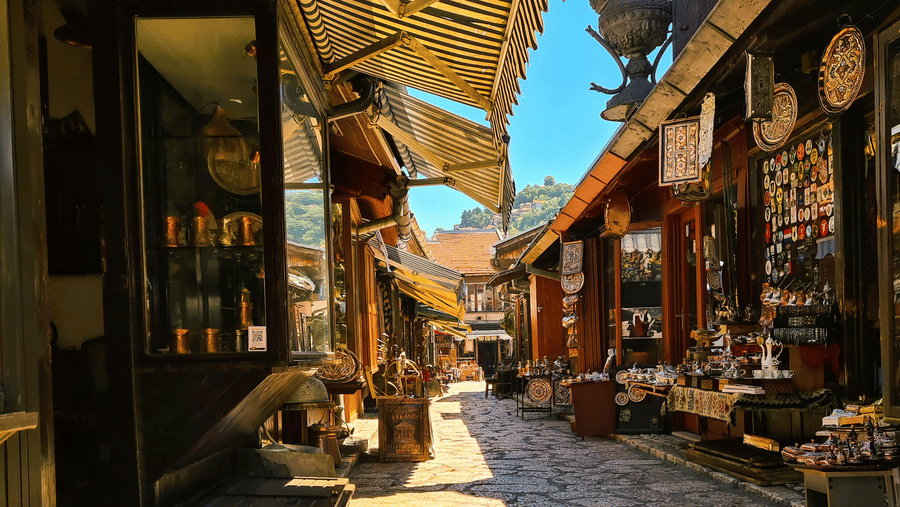 A narrow sunlit alleyway lined with small shops featuring yellow striped awnings and various goods displayed outside.