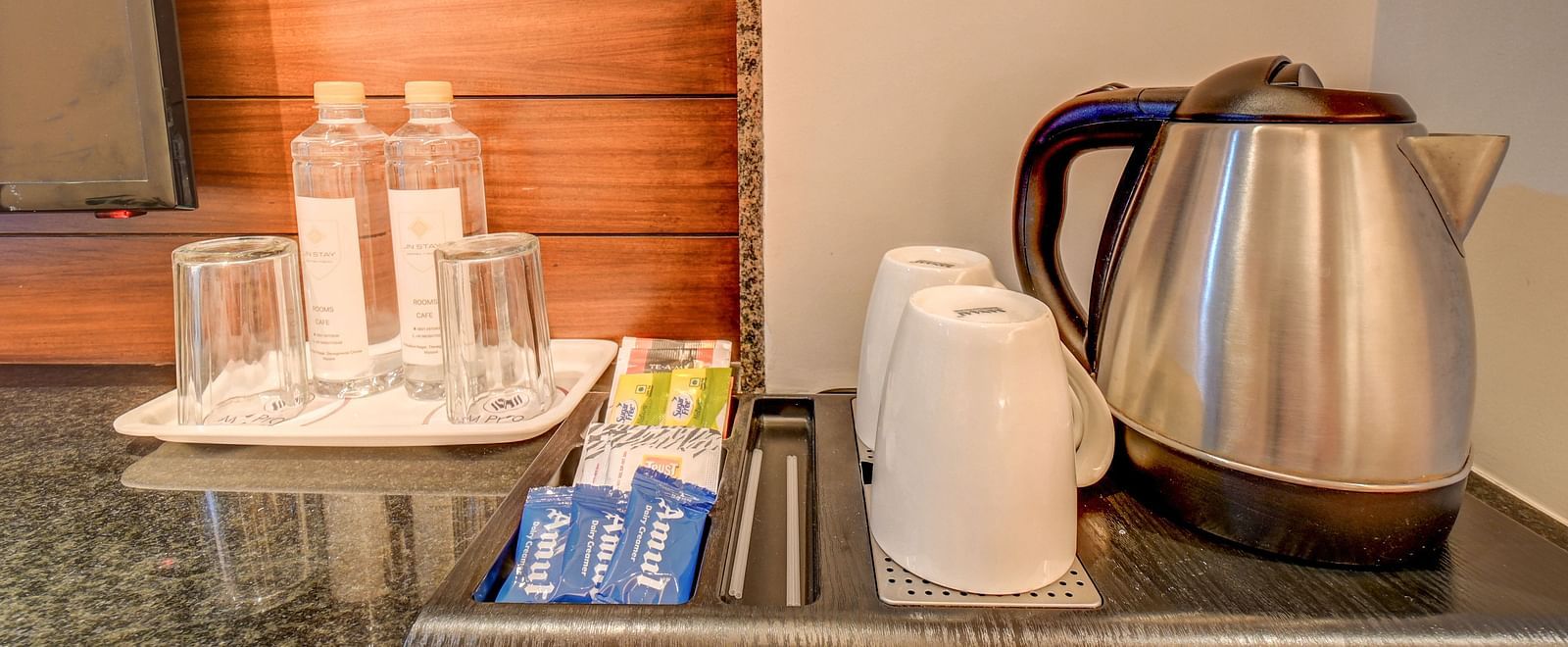 Two trays at Harigovinda's JNStay: one with glasses and water bottles, and the other with cups, snacks, and a kettle