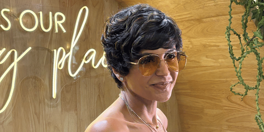 A side angle of woman with a short pixie haircut and tinted sunglasses wearing a white strapless top with black polka dots in front of a wooden wall with neon signage.