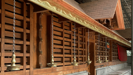 A wooden slatted wall structure with hanging metal oil lamps sits above a stone ledge under a covered roof