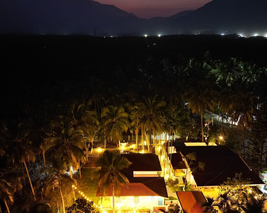 Aerial view of a resort at dusk with illuminated buildings and palm trees, set against a backdrop of mountains.