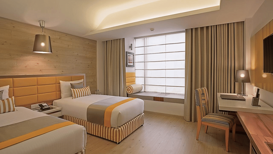 An Deluxe Twin Room at Barsana Boutique Hotel, Kolkata, featuring twin beds draped in matching linens, a large window, and a workspace with a chair.