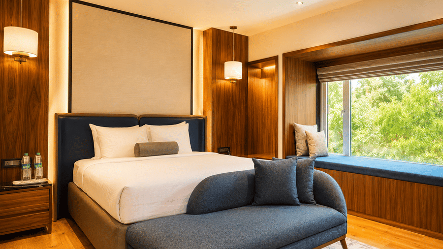 Bedroom at Pride Premier Pune with a king size bed, blue bench and large window overlooking greenery.