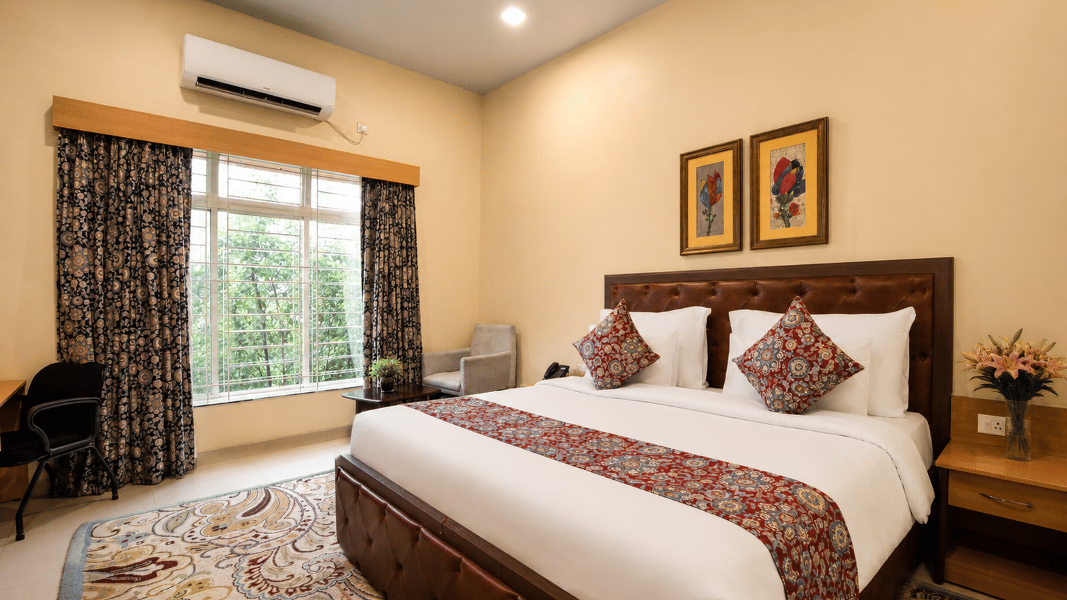 Deluxe room with a king bed, leather headboard, patterned runner, work desk, armchair and a window overlooking greenery at MAYFAIR World Cup Village, Rourkela.