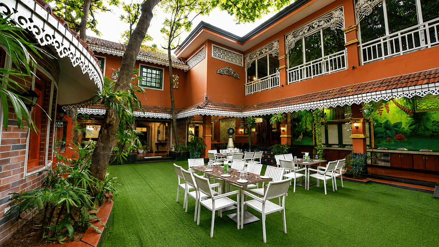 Aangan courtyard restaurant with white tables and chairs on a lawn, terracotta building with fretwork trim and a painted mural at MAYFAIR Garden, Rourkela.