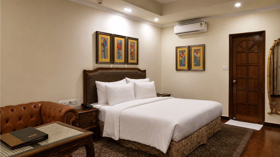 Deluxe king room with white bed linen, upholstered headboard, framed floral prints, leather sofa, carved desk and a patterned rug at MAYFAIR Garden, Rourkela.