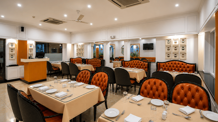 Tandoor restaurant with orange tufted banquettes, tables laid with cream cloths, framed butterfly displays and a marble floor at MAYFAIR Garden, Rourkela.