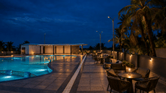 Wide dusk view of the swimming pool at Pride Elite Mandarmani with an illuminated curved pool, lounge seating on the deck, palm trees, and a poolside pavilion.