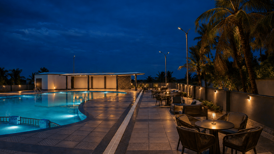 Wide dusk view of the swimming pool at Pride Elite Mandarmani with an illuminated curved pool, lounge seating on the deck, palm trees, and a poolside pavilion.