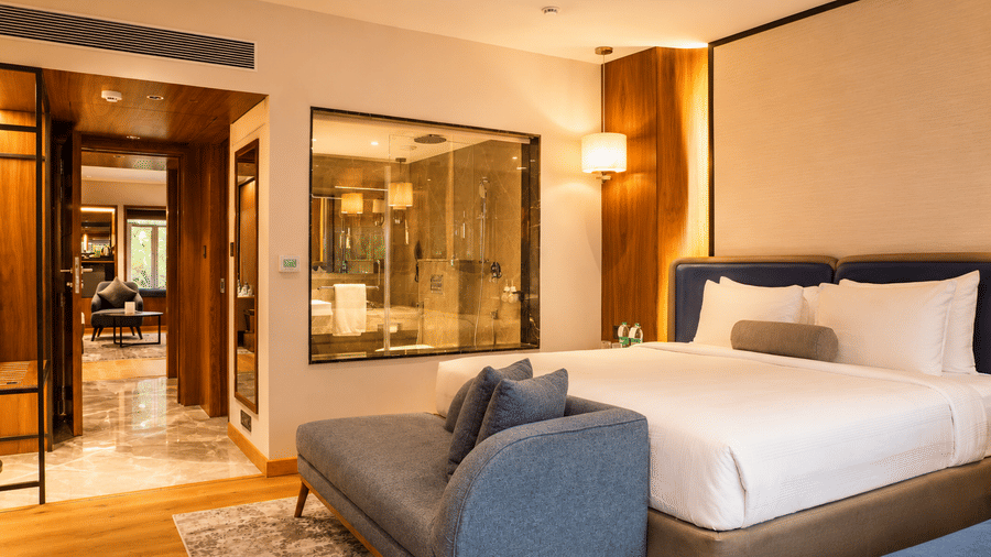 Guest room at Pride Premier Pune featuring a king-size bed, blue armchair and open layout with warm lighting.