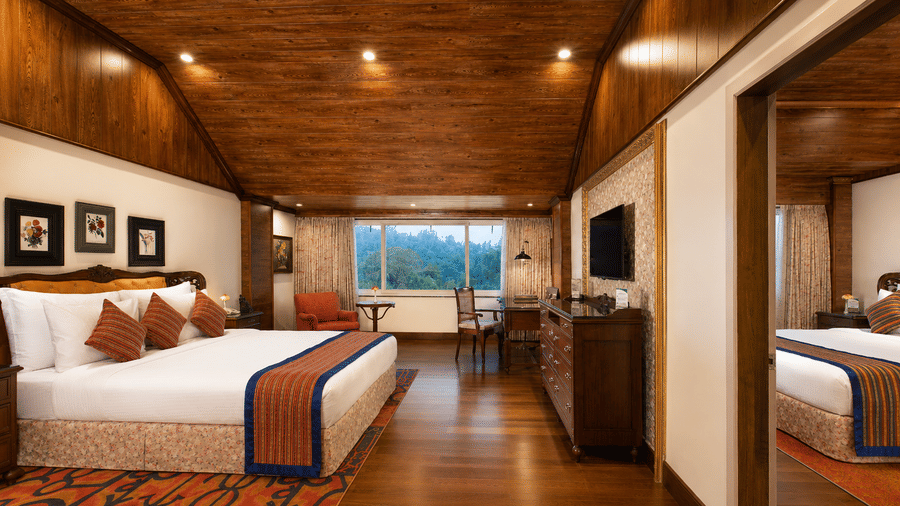 Family Room at MAYFAIR Himalayan Spa Resort Kalimpong. A bed with pillows faces a television on a cabinet. Wood panelling covers the ceiling and floor. A window at the far end looks out towards trees. A mirror reflects a portion of the room.