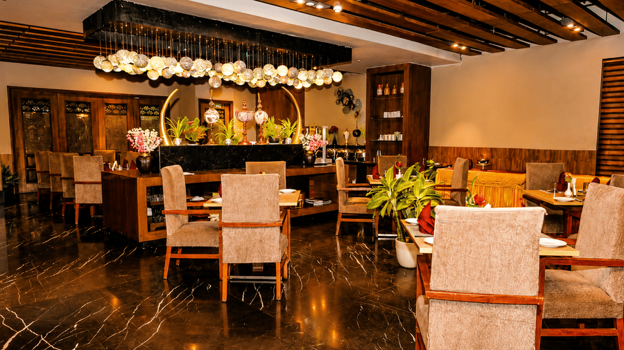 Restaurant dining area with round tables, upholstered chairs, a floral display, exposed wooden beams overhead and dark marble-effect flooring.