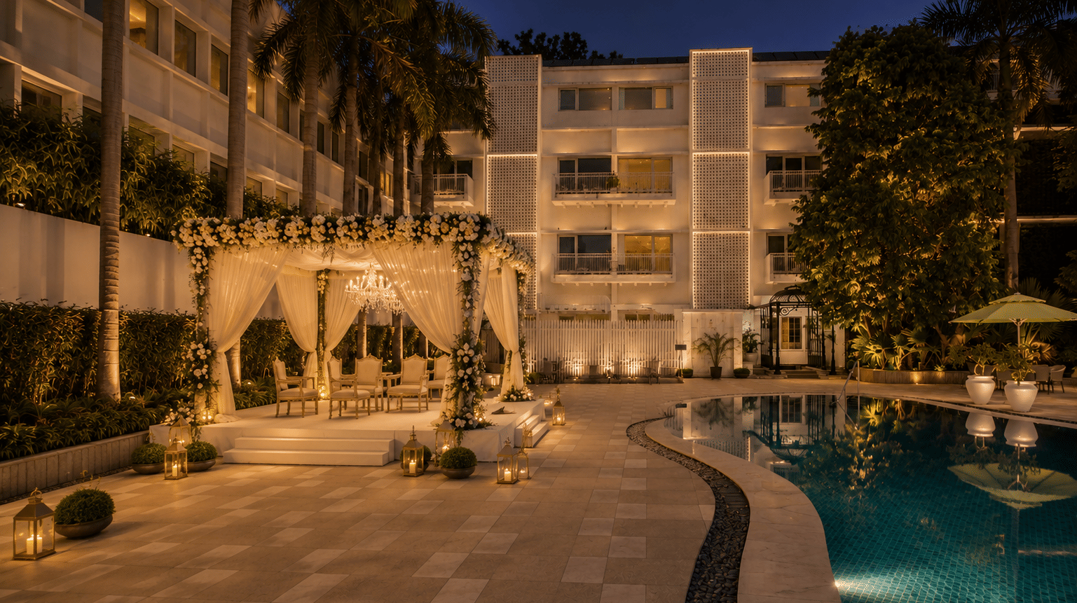 Night view of an illuminated outdoor cabana with white drapes set beside a swimming pool at a hotel courtyard - The Claridges New Delhi