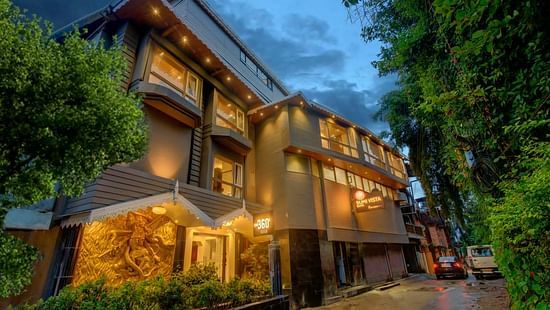 An image of a well lit facade surrounded by trees and there is also a driveway near the entrance - Sumi Yashshree Hotels & Resorts