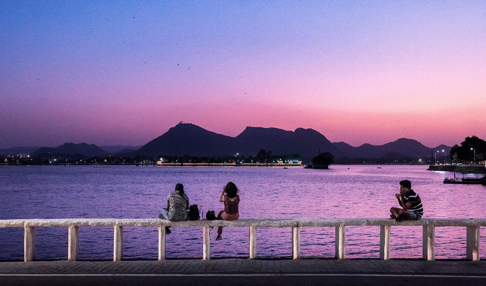 Three people sit on a railing overlooking a lake at dusk.